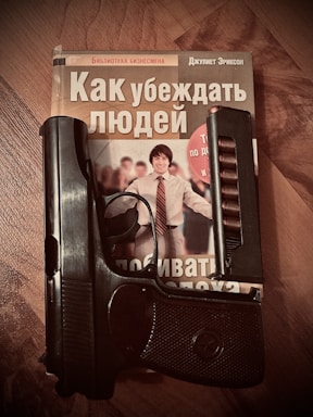 A book with a cover featuring a smiling person is placed on a wooden surface. A handgun is resting on the book, partially covering the title and image.