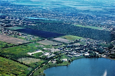An aerial view of a landscape featuring a large expanse of green fields and forested areas. Residential buildings and roads are scattered throughout the landscape, showing urban development. A large body of water is visible in the lower part of the image, possibly a lake or river. The horizon showcases a vast, flat terrain extending into the distance with more urban areas.
