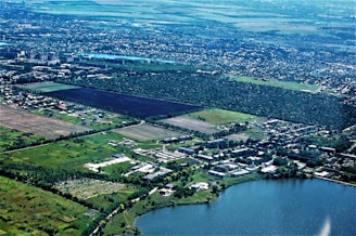 An aerial view of a landscape featuring a large expanse of green fields and forested areas. Residential buildings and roads are scattered throughout the landscape, showing urban development. A large body of water is visible in the lower part of the image, possibly a lake or river. The horizon showcases a vast, flat terrain extending into the distance with more urban areas.