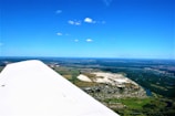 Aerial view of a landscape with a visible airplane wing in the foreground. The scenery includes a mix of fields, small bodies of water, and clusters of buildings. There are also some distinct white rock formations in the center portion of the image. The sky is clear with minimal clouds, and there is a vast horizon visible.