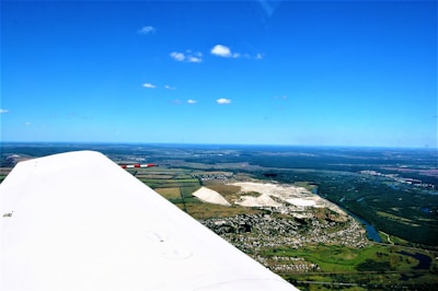 Aerial view of a landscape with a visible airplane wing in the foreground. The scenery includes a mix of fields, small bodies of water, and clusters of buildings. There are also some distinct white rock formations in the center portion of the image. The sky is clear with minimal clouds, and there is a vast horizon visible.