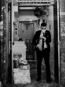 A man wearing a suit and a face mask stands at the entrance of a building. There are boxes and bags piled on the ground next to the open door. The building's interior appears to be a narrow space with a fan attached to the wall and signage in an Asian script visible. A person is seated at a desk partially visible in the background.