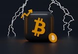 A 3D rendering features a black square with an orange Bitcoin symbol and an accompanying small coin with the same symbol. Behind, white lightning bolts illuminate the dark background, adding a dynamic visual element.