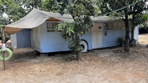 An old bus is repurposed into a shelter, partially covered by a makeshift awning. It is parked under the shade of trees, with branches and leaves surrounding it. A small path of stone tiles leads to the entrance, while a detached green hanging tire can be seen nearby. The ground around is dry and sandy, suggesting a rustic or possibly rural setting.