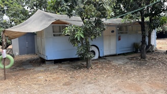 An old bus is repurposed into a shelter, partially covered by a makeshift awning. It is parked under the shade of trees, with branches and leaves surrounding it. A small path of stone tiles leads to the entrance, while a detached green hanging tire can be seen nearby. The ground around is dry and sandy, suggesting a rustic or possibly rural setting.
