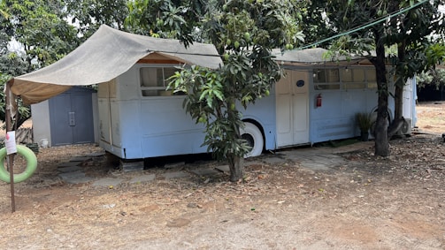 An old bus is repurposed into a shelter, partially covered by a makeshift awning. It is parked under the shade of trees, with branches and leaves surrounding it. A small path of stone tiles leads to the entrance, while a detached green hanging tire can be seen nearby. The ground around is dry and sandy, suggesting a rustic or possibly rural setting.
