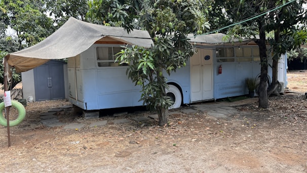 An old bus is repurposed into a shelter, partially covered by a makeshift awning. It is parked under the shade of trees, with branches and leaves surrounding it. A small path of stone tiles leads to the entrance, while a detached green hanging tire can be seen nearby. The ground around is dry and sandy, suggesting a rustic or possibly rural setting.