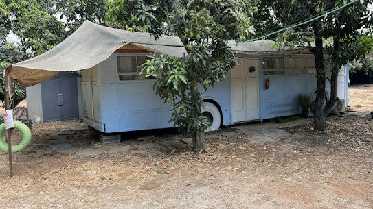 An old bus is repurposed into a shelter, partially covered by a makeshift awning. It is parked under the shade of trees, with branches and leaves surrounding it. A small path of stone tiles leads to the entrance, while a detached green hanging tire can be seen nearby. The ground around is dry and sandy, suggesting a rustic or possibly rural setting.