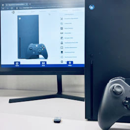A computer monitor displays a webpage featuring an Xbox console and controller, with various repair options listed on the right. In front of the monitor, there is an actual Xbox console and a controller positioned on a white surface. A small USB device is placed between the console and the monitor.