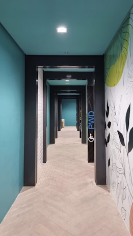A corridor with a series of doorways, painted in a teal color with contrasting black and white elements. The right wall features a decorative mural with leaves, adding a touch of nature to the modern design. Soft light comes from ceiling fixtures, illuminating the wood-patterned flooring, creating a clean and contemporary space.