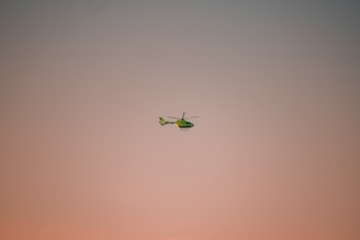 A green helicopter is flying in the sky, which is painted with soft hues of pink and peach during either sunrise or sunset. The helicopter is slightly tilted forward and appears to be in motion.