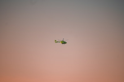 A green helicopter is flying in the sky, which is painted with soft hues of pink and peach during either sunrise or sunset. The helicopter is slightly tilted forward and appears to be in motion.