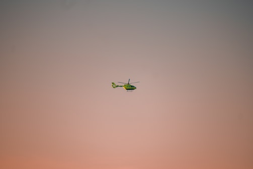 A green helicopter is flying in the sky, which is painted with soft hues of pink and peach during either sunrise or sunset. The helicopter is slightly tilted forward and appears to be in motion.
