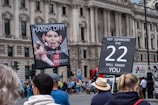 A group of people are gathered in front of a historic building with large columns. Two prominent signs are held up by individuals. The first sign features an image of a caricature with the words 'HANDS OFF! OUR RIGHT TO PROTEST!'. The second sign reads 'HEY JOHNSON! THE 22 WILL FINISH YOU'. The crowd appears to be participating in a protest or demonstration.