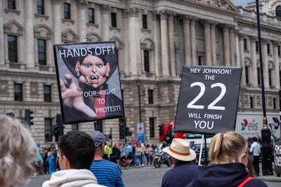 A group of people are gathered in front of a historic building with large columns. Two prominent signs are held up by individuals. The first sign features an image of a caricature with the words 'HANDS OFF! OUR RIGHT TO PROTEST!'. The second sign reads 'HEY JOHNSON! THE 22 WILL FINISH YOU'. The crowd appears to be participating in a protest or demonstration.