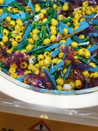 A collection of small, colorful plastic toy pieces consisting primarily of yellow heads with facial expressions, green and blue accessories, and some transparent purple butterfly-like pieces mixed together.