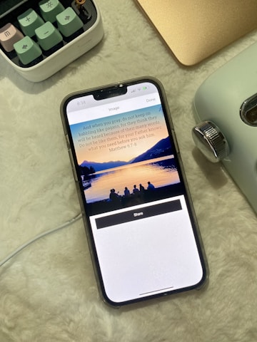 A smartphone displaying a Bible verse and a serene image of a sunset over a lake with silhouetted people. It rests on a soft, light-colored surface alongside a retro camera and part of a keyboard.
