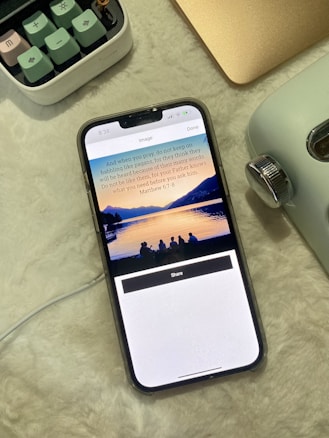 A smartphone displaying a Bible verse and a serene image of a sunset over a lake with silhouetted people. It rests on a soft, light-colored surface alongside a retro camera and part of a keyboard.