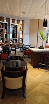 A modern and elegant interior space, featuring wooden furniture and vibrant decorative elements. The room contains tables and chairs arranged neatly, with shelves displaying ornamental vases and artwork in the background. The lighting is warm, creating a cozy and inviting atmosphere. A small plant on the table adds a touch of greenery.