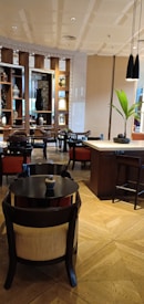 A modern and elegant interior space, featuring wooden furniture and vibrant decorative elements. The room contains tables and chairs arranged neatly, with shelves displaying ornamental vases and artwork in the background. The lighting is warm, creating a cozy and inviting atmosphere. A small plant on the table adds a touch of greenery.