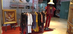 A display of traditional clothing with various garments hanging on a rack, including white, black, yellow, and purple outfits. A mannequin dressed in red traditional attire with a large straw hat stands beside the rack. To the left, a framed emblem featuring a gold eagle is propped up on an easel. Behind the display, there are glass cases and promotional banners.