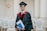 A person wearing a graduation gown and cap is holding a diploma. They are standing in a room with white columns and empty rows of chairs. The gown is black with red and gold accents.