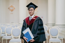 A person wearing a graduation gown and cap is holding a diploma. They are standing in a room with white columns and empty rows of chairs. The gown is black with red and gold accents.
