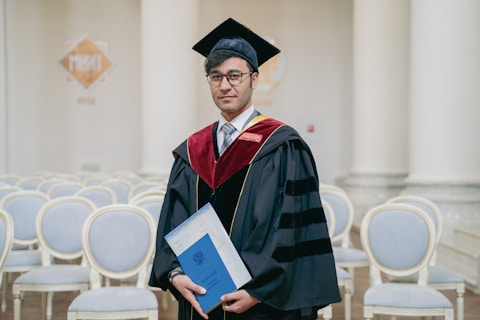 A person wearing a graduation gown and cap is holding a diploma. They are standing in a room with white columns and empty rows of chairs. The gown is black with red and gold accents.