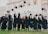 A group of graduates in black gowns with maroon accents joyfully throwing their caps into the air in front of an ornate building. The building has large arched windows and decorative architectural details.