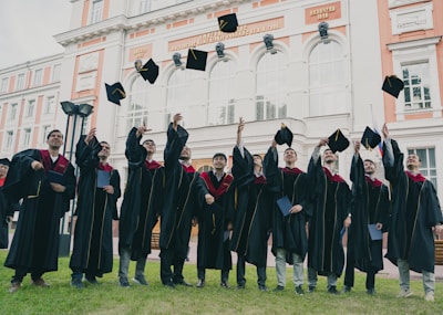 A group of graduates in black gowns with maroon accents joyfully throwing their caps into the air in front of an ornate building. The building has large arched windows and decorative architectural details.
