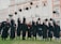 A group of graduates in black gowns with maroon accents joyfully throwing their caps into the air in front of an ornate building. The building has large arched windows and decorative architectural details.