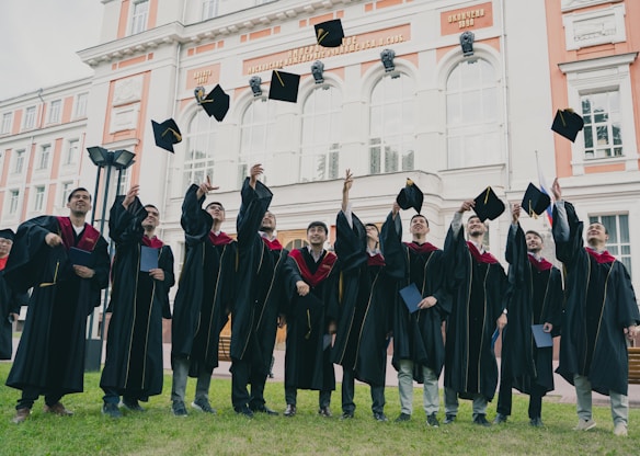 A group of graduates in black gowns with maroon accents joyfully throwing their caps into the air in front of an ornate building. The building has large arched windows and decorative architectural details.