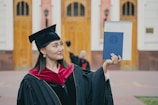 A graduate proudly holds up a diploma while wearing a traditional cap and gown. The setting appears to be outside a formal building with wooden doors and pillars visible in the background. The graduate is smiling slightly, exuding a sense of accomplishment.