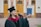 A graduate proudly holds up a diploma while wearing a traditional cap and gown. The setting appears to be outside a formal building with wooden doors and pillars visible in the background. The graduate is smiling slightly, exuding a sense of accomplishment.