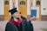 A graduate proudly holds up a diploma while wearing a traditional cap and gown. The setting appears to be outside a formal building with wooden doors and pillars visible in the background. The graduate is smiling slightly, exuding a sense of accomplishment.