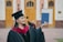 A graduate proudly holds up a diploma while wearing a traditional cap and gown. The setting appears to be outside a formal building with wooden doors and pillars visible in the background. The graduate is smiling slightly, exuding a sense of accomplishment.