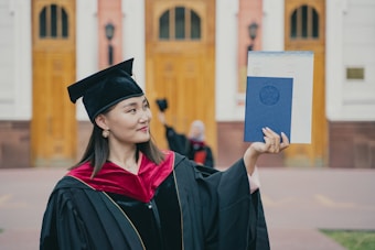 A graduate proudly holds up a diploma while wearing a traditional cap and gown. The setting appears to be outside a formal building with wooden doors and pillars visible in the background. The graduate is smiling slightly, exuding a sense of accomplishment.