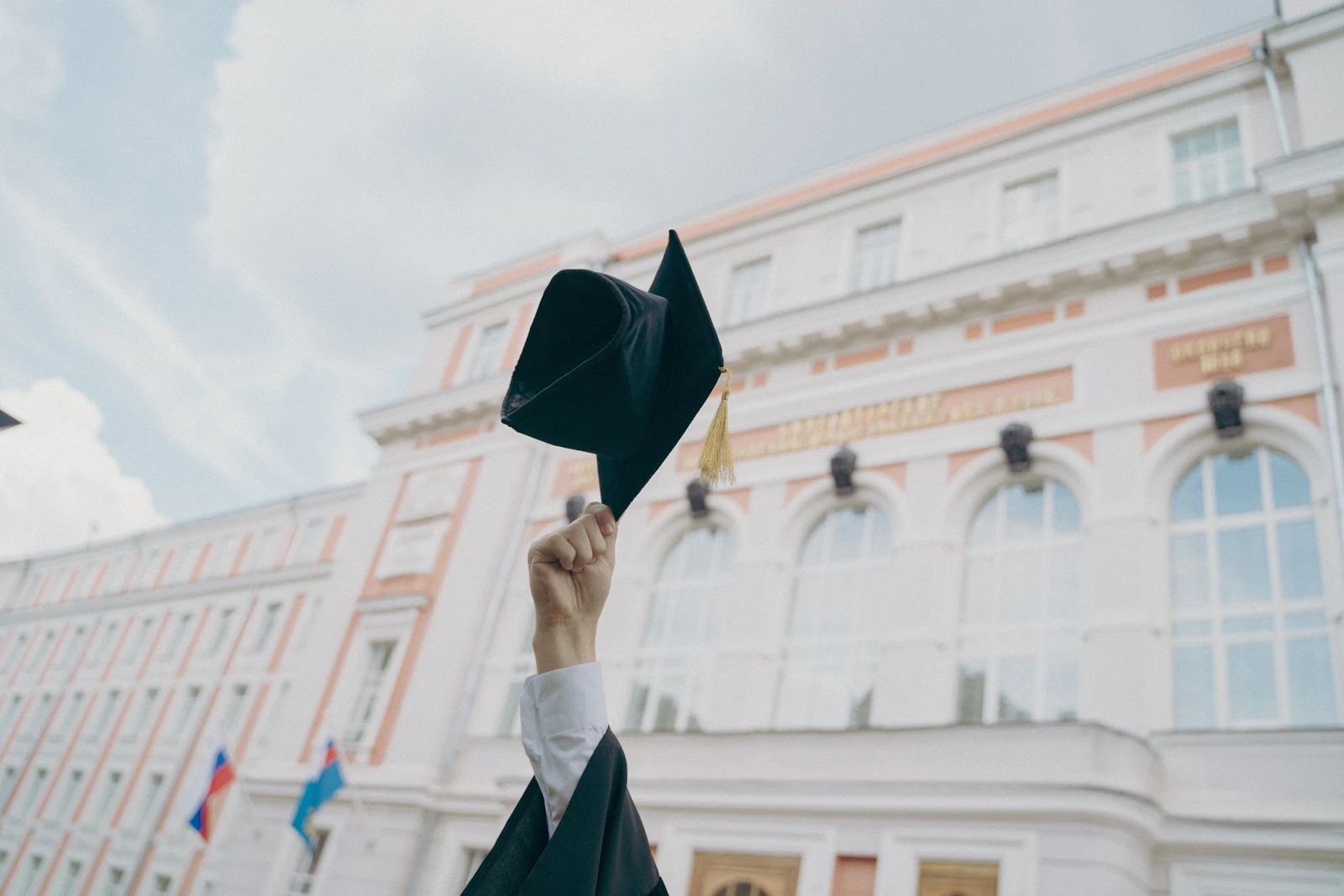 A hand holding a black graduation cap up in front of a large university building