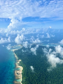 Aerial view of an expansive island covered in dense green forest surrounded by clear blue ocean waters. White sandy beaches line the coast, and fluffy clouds are scattered across the bright blue sky.