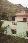 A house with white walls and red-tiled roof is situated amidst lush green foliage. The structure features balconies with white railings and windows with red curtains. In the background, there are rolling hills covered in greenery, suggesting a mountainous location.
