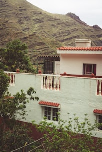 A house with white walls and red-tiled roof is situated amidst lush green foliage. The structure features balconies with white railings and windows with red curtains. In the background, there are rolling hills covered in greenery, suggesting a mountainous location.