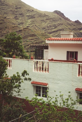 A house with white walls and red-tiled roof is situated amidst lush green foliage. The structure features balconies with white railings and windows with red curtains. In the background, there are rolling hills covered in greenery, suggesting a mountainous location.