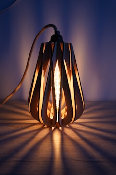 A decorative light fixture with a modern design, featuring a filament bulb encased in a wooden frame with elongated cutout patterns. The light casts intricate shadow patterns on the surface below, creating a warm and inviting ambiance against a muted blue background.