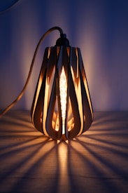 A decorative light fixture with a modern design, featuring a filament bulb encased in a wooden frame with elongated cutout patterns. The light casts intricate shadow patterns on the surface below, creating a warm and inviting ambiance against a muted blue background.