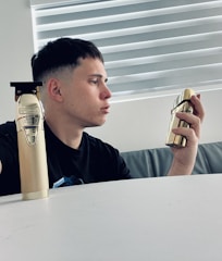 A person with short dark hair is sitting at a white table, holding a gold-colored perfume bottle in their right hand. A gold-colored hair trimmer is placed on the table. The background features a window with horizontal blinds.