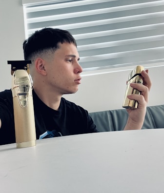 A person with short dark hair is sitting at a white table, holding a gold-colored perfume bottle in their right hand. A gold-colored hair trimmer is placed on the table. The background features a window with horizontal blinds.