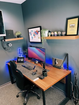 A well-organized home office or gaming setup with a wooden desk, a curved monitor, a keyboard, and a mouse. There is a digital clock displaying 17:53 on the desk. Shelves mounted on the wall hold several small plants, decorative figures, and framed pictures, with one frame displaying a 'Do What You Love' quote. The lighting includes blue LED strips under the desk and around the room. A black gaming chair is positioned in front of the desk, and a webcam is mounted on the wall. The walls are painted in a dark gray shade.