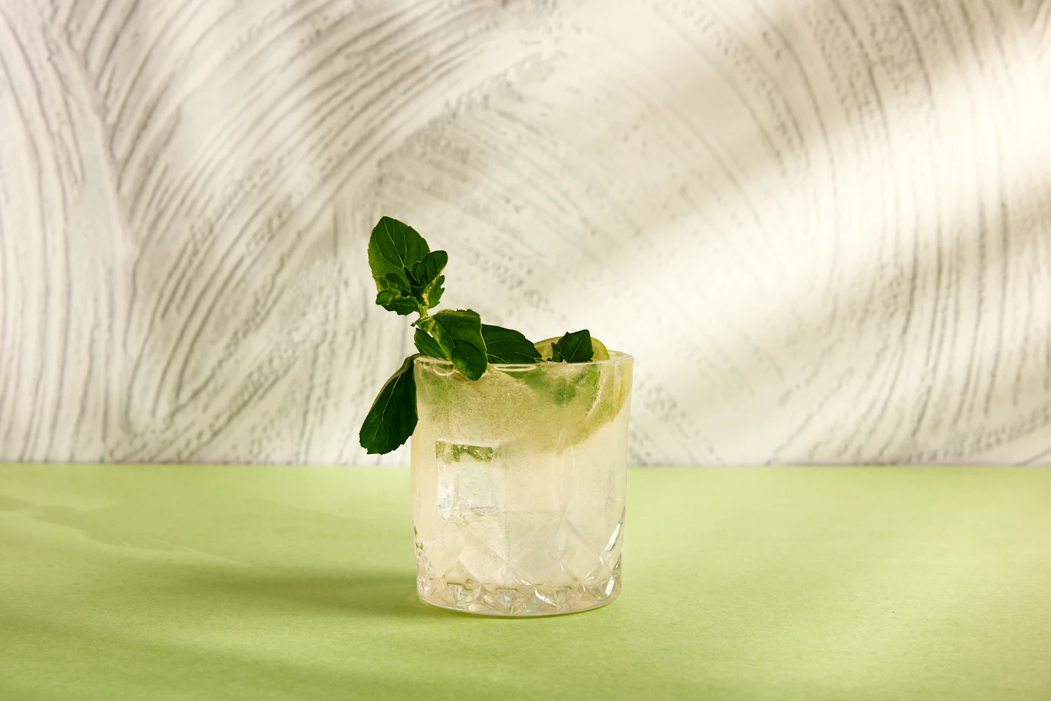 A gin and tonic with lime and a mint sprig on a pale green table