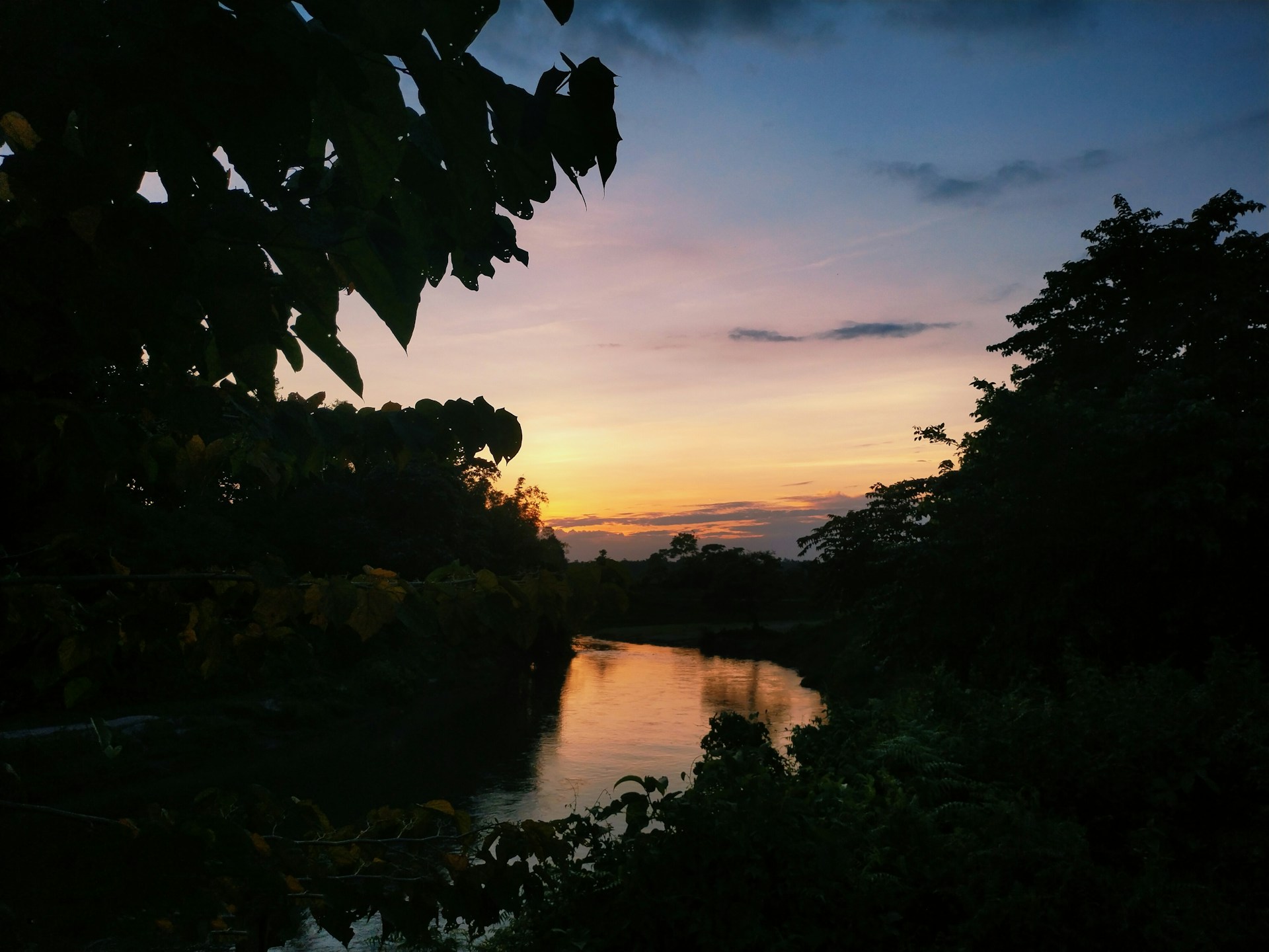 A stunning sunset over a calm river reflecting the colors of the sky, framed by natural foliage.