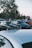 A group of parked cars is seen in an outdoor setting typical of a car meet. People are gathered around, some standing near the cars and conversing. The vehicles are older models, with visible details like decals and custom features. Trees and a clear sky form the backdrop, creating a relaxed and social atmosphere.